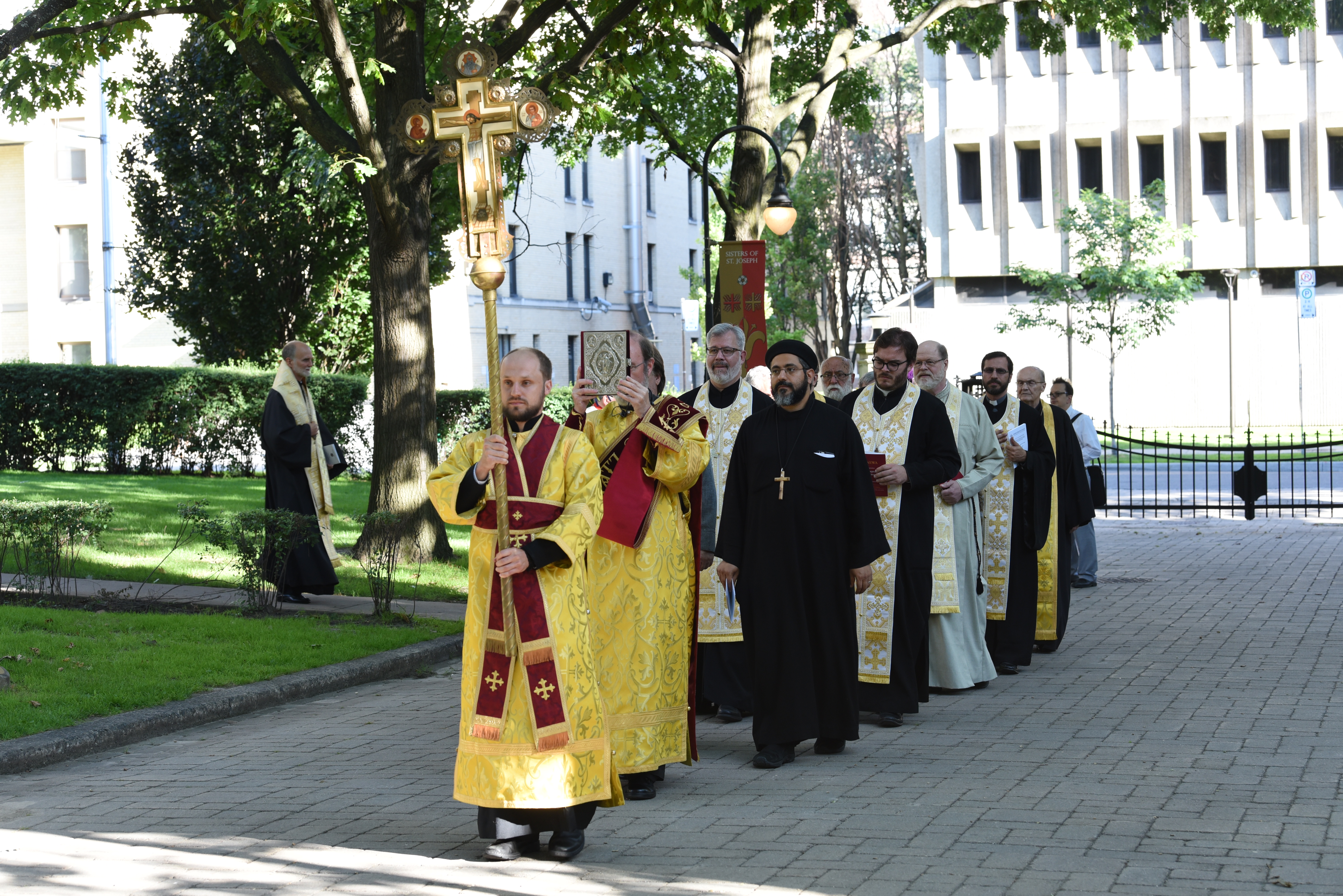 The image depicts various religious people walking calmly in a line with the lead person holding a large golden scepter.