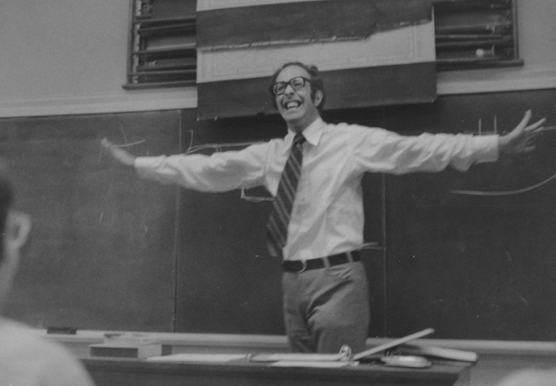 Henri J.M. Nouwen in a classroom in front of chalk board, arms stretched out with a big smile.