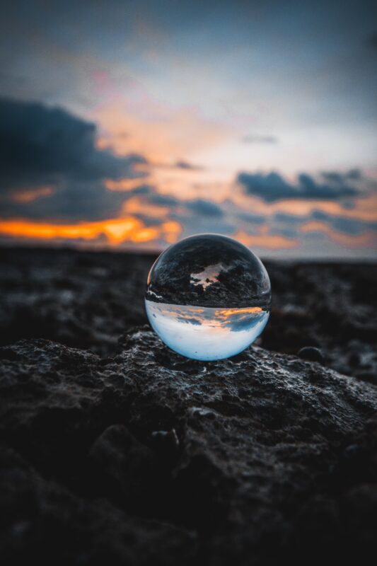 Image depicts a glass marble on a mound of dirt. The marble gives an upside-down reflection of the sky and ground in the background. 
