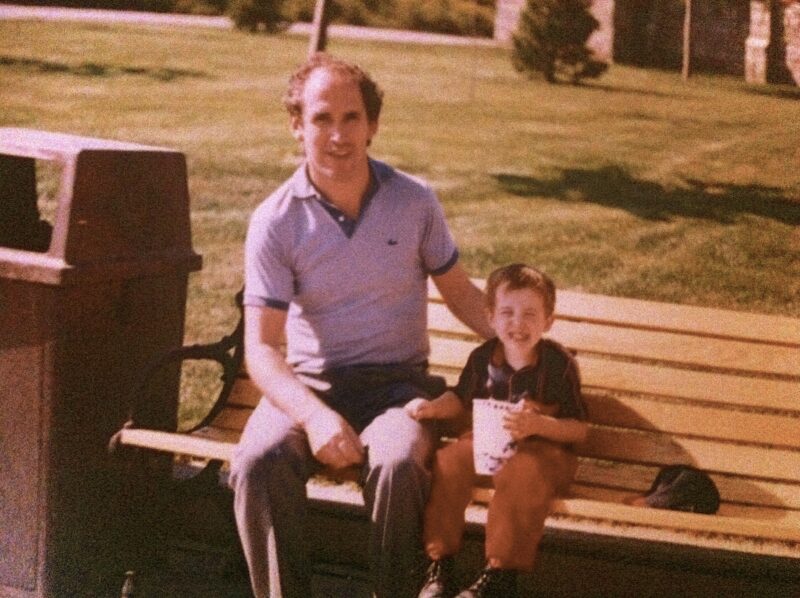 Vintage image depicts a man sitting next to his young son on a park bench. The son is holding a large drink in a takeout cup. 