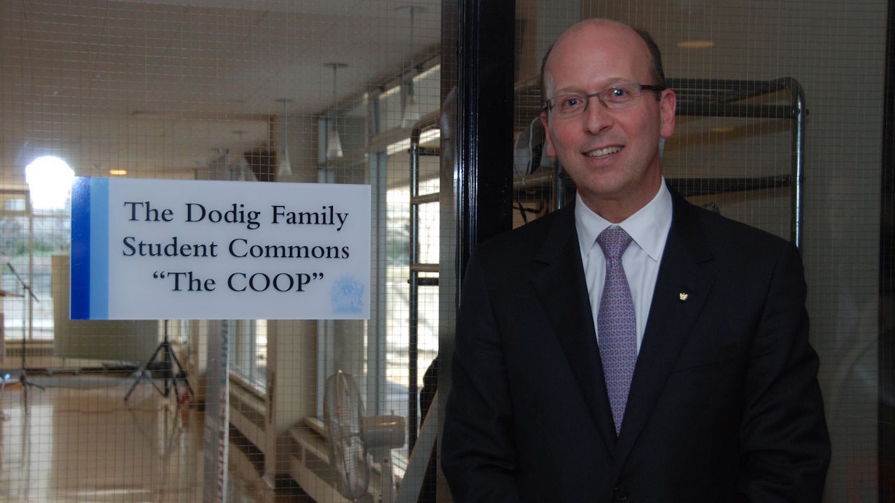 Image depicts alumnus Victor Dodig standing next to a sign for the Dodig Family Student Commons (known as the Coop) in Brennan Hall on the St. Michael's campus. 