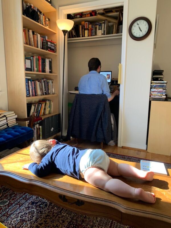 A toddler rests face-down on a coffee table facing a man working at a desk in a closet. 