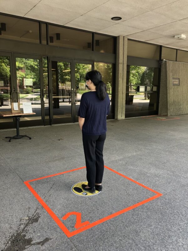 Image depicts a woman standing in an area marked with tape to remain at an appropriate social distance while waiting to claim her curbside pickup request at the Kelly Library