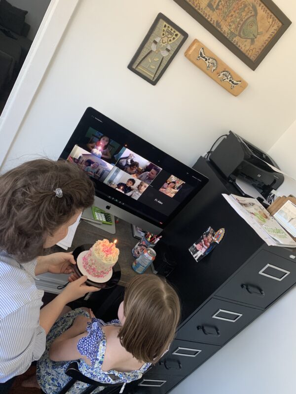 A woman and child place a small birthday cake with a candle on top on a desk in front of a computer, where a video call is in progress