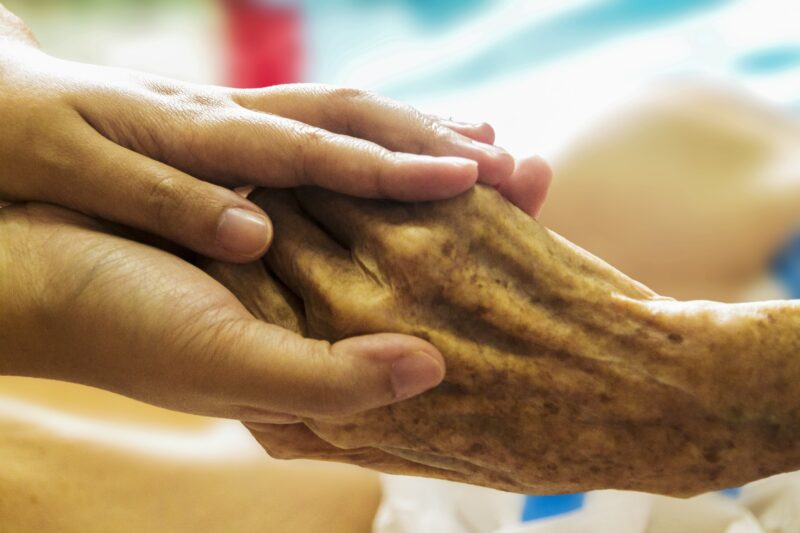 A hospice caregiver clasps the hands of an elderly patient 