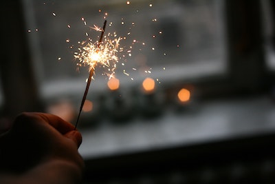A lit sparkler held in front of a window and a row of candles 