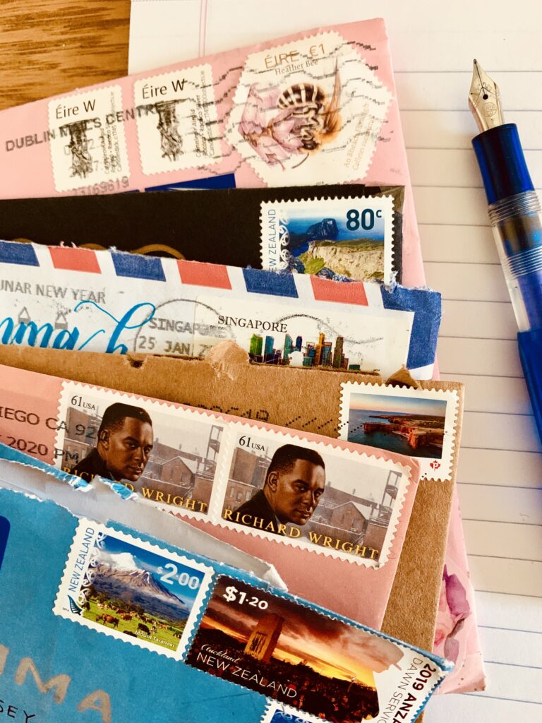 Close focus photograph of six envelopes resting on a pad of paper next to a fountain pen. The envelopes have stamps from Ireland, New Zealand, Singapore, Canada, and USA.