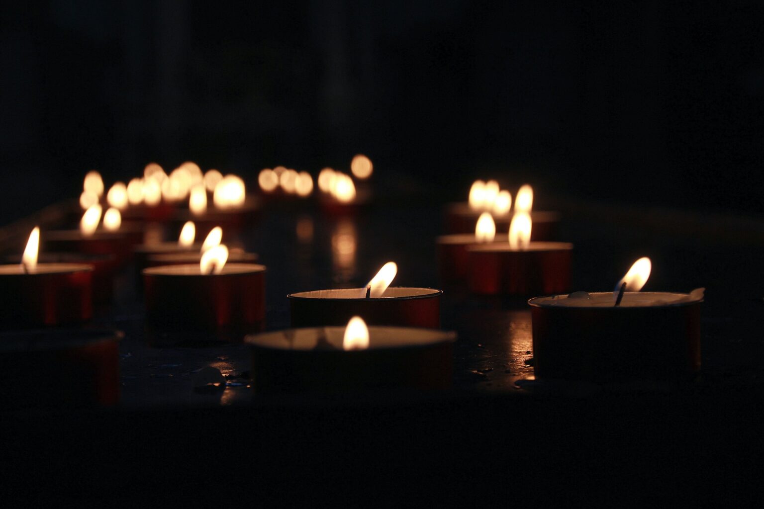 Photograph of multiple tea light candles floating on a dark body of water at night. There are many more candle flames out of focus in the background.