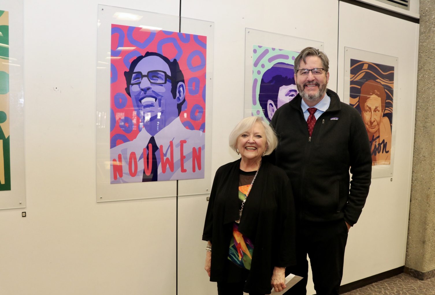 Photograph of Karen Pascal and David Sylvester, smiling, in front of an illustration of Henri Nouwen on a Kelly Library wall.