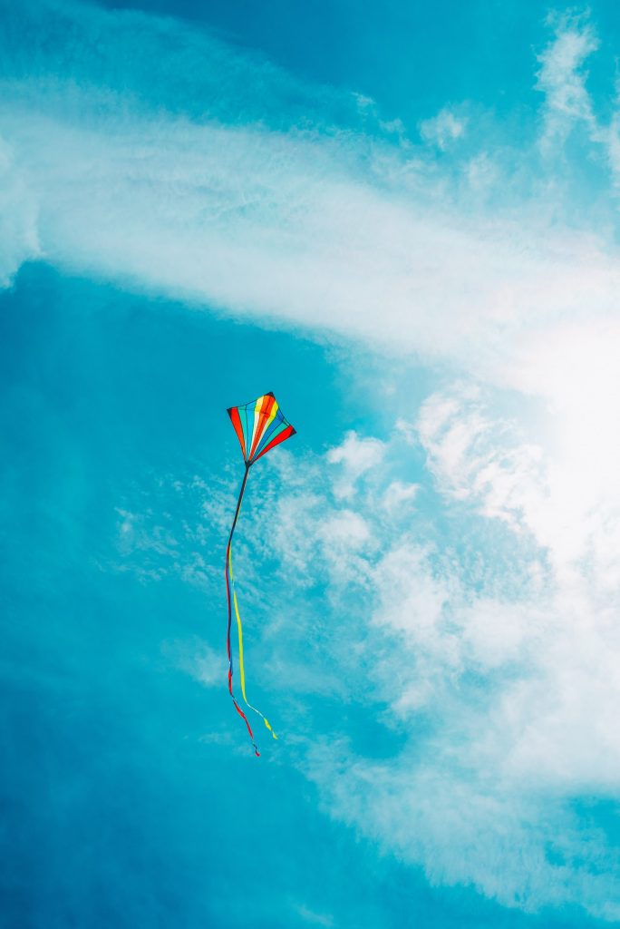 Photograph of a colourful kite with a long tail flying against the background of a bright blue sky and wispy clouds.