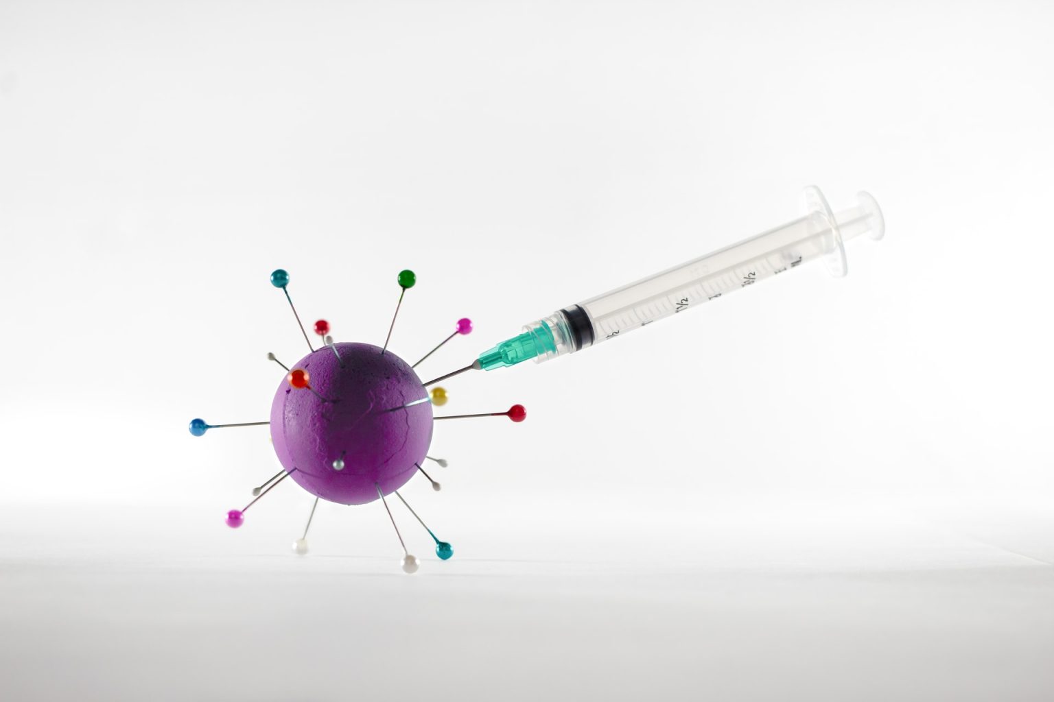 Photograph of a syringe with hypodermic needle stuck into an artistic representation of the coronavirus molecule (a painted styrofoam ball with coloured pins stuck in it).