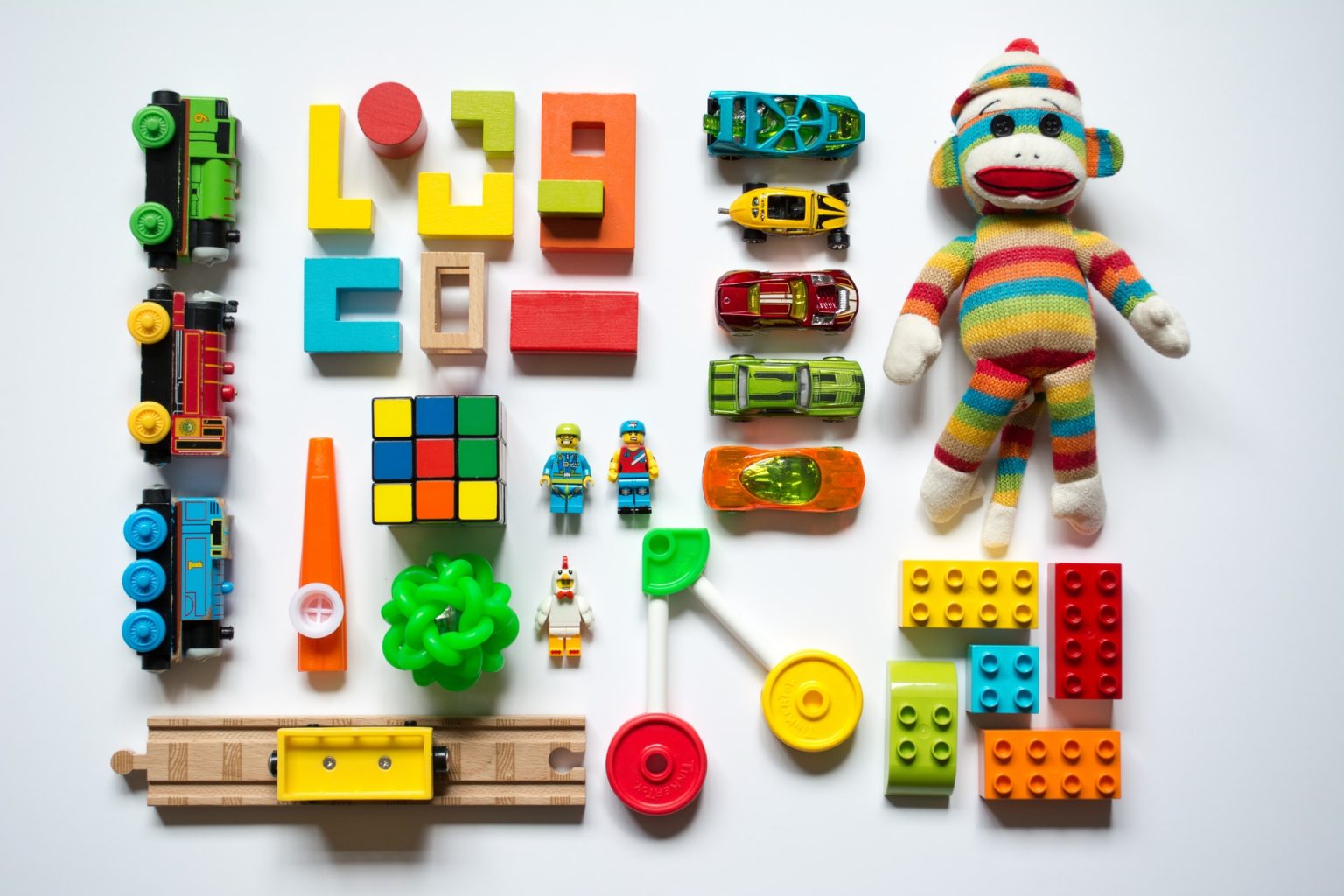 Still life photograph of a white background laid out with multiple brightly coloured children's toys: Thomas trains, a train track, blocks, a Rubik's cube, a kazoo, a bouncing ball, Lego figures, Hot Wheels, a striped sock monkey, Lego blocks, and connecting toys.