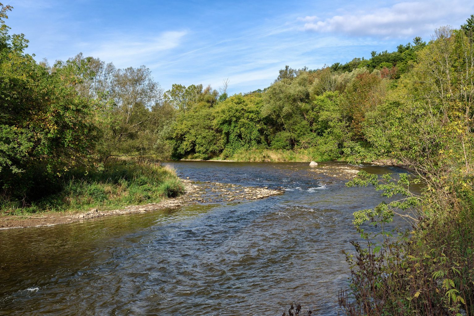 Photograph of a curve in the Credit River, Mississauga, Ontario. There are green trees in the midground and a bright blue sky in the background.