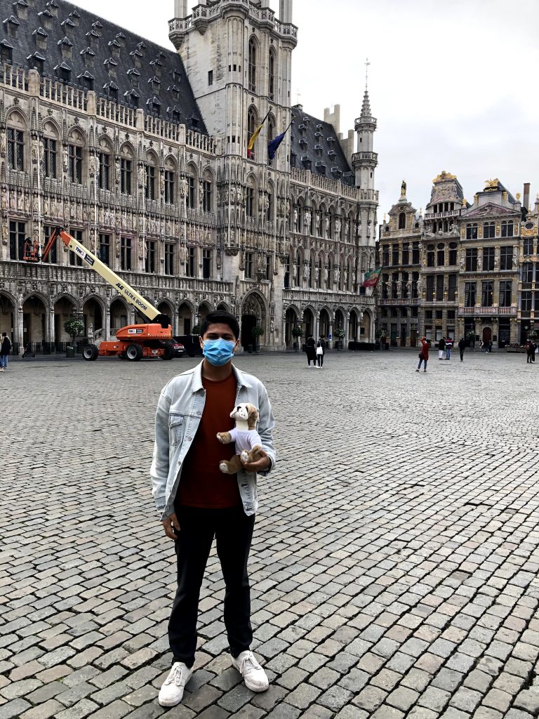 Photograph of post author Nicolas Vergara Ruiz standing in front of The Grand Place, the central square of Brussels, Belgium, wearing a blue medical mask and holding a Basil the bulldog plushie.