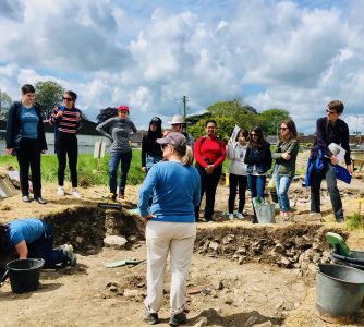 A woman stands in a shallow archeological pit outdoors with students standing above its edge looking in at the work being done there