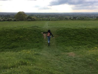 A woman walks up a path through grassy fields outdoors towards trees and fields far away 