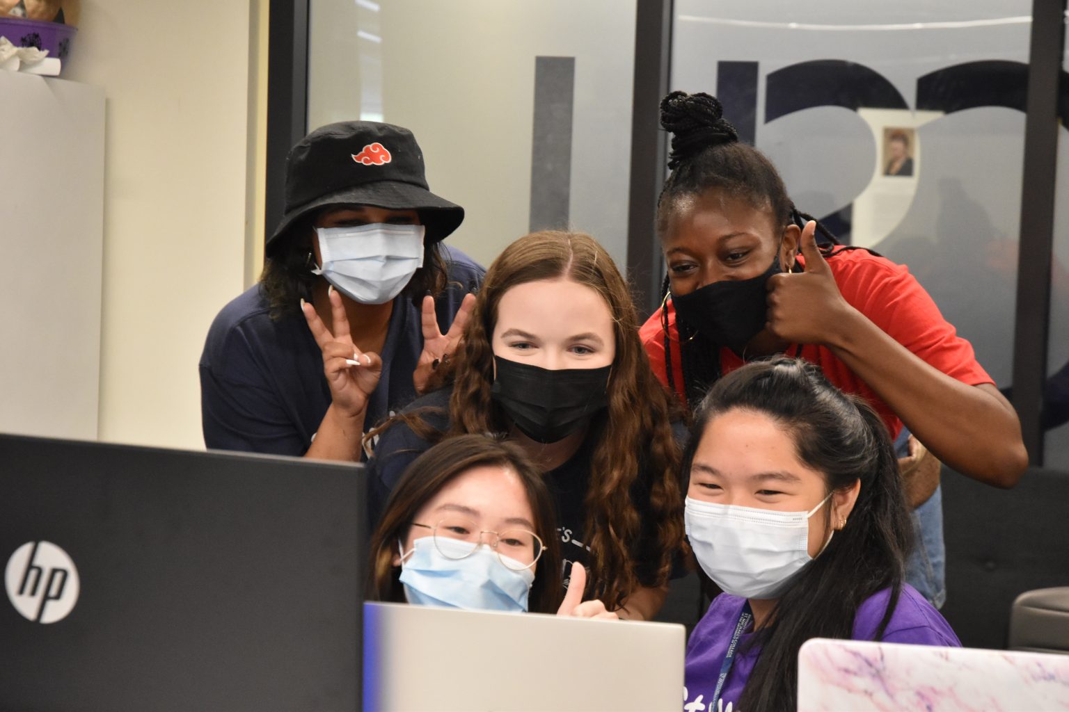 Photograph of five St. Mike's orientation leaders behind three monitor screens, wearing masks, smiling with their eyes, and offering thumbs ups and peace signs for the computer cameras.