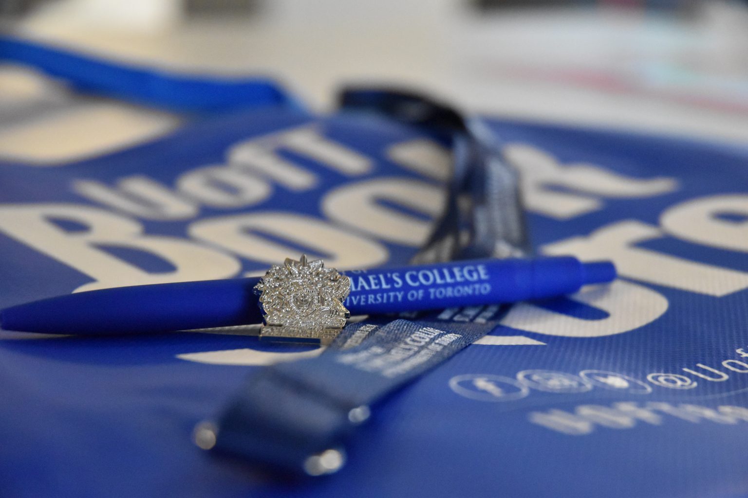 Close focus photograph of a small silver pin shaped like the University of St. Michael's College crest, a pen with the USMC logo, and a  blue lanyard all sitting on a U of T Book Store tote bag.