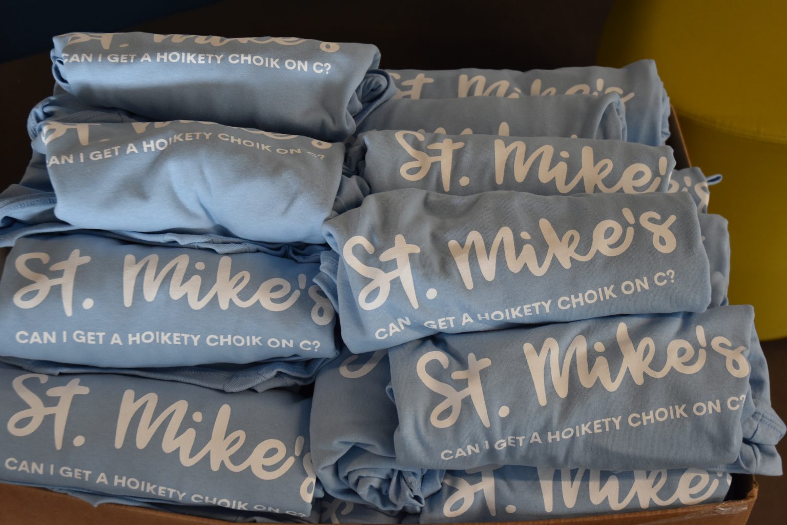 Photograph of two stacks of light blue tshirts with the text "St. Mike's" in large script font and "Can I get a Hoikety Choik on C?" written below. 