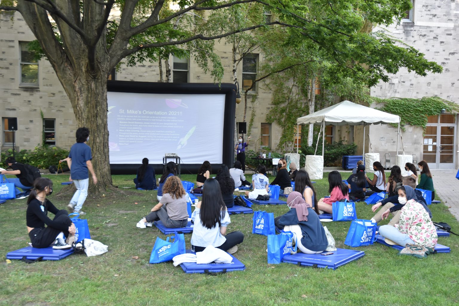 Photograph of a number of Class of 2025 students seated on blue mats on the St. Mike's quad lawn, spaced 6 feet apart, watching a presentation on a large outdoor screen.