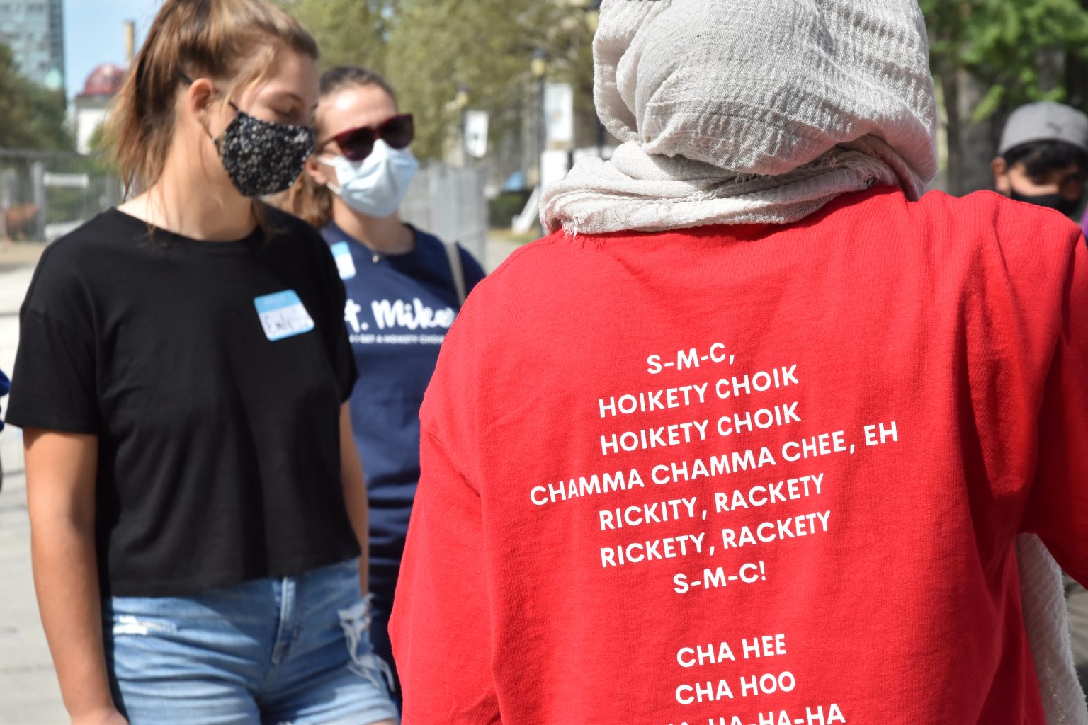 Close up image of a red orientation leader shirt, worn by a leader wearing a headscarf, with the following text visible: "S-M-C, Hoikety Choik Hoikety Choik Chamma Chamma Chee, Eh Rickity, Rackety Rickity, Rackety S-M-C! Cha Hee Cha Hoo…"