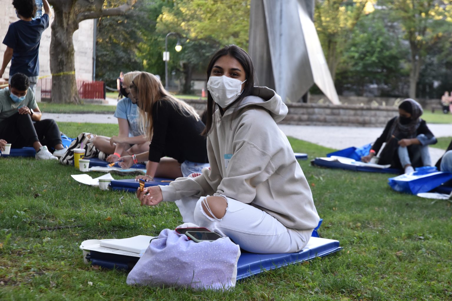 Photograph of a new St. Mike's student wearing a mask and smiling with their eyes, holding up a paintbrush while sitting on the St. Mike's quad