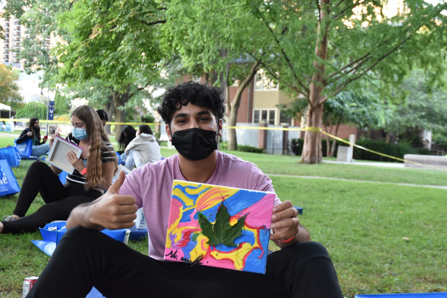 Photograph of a new St. Mike's student holding up a colourful abstract painting with a real leaf placed on the center. He is giving the camera a thumbs up.