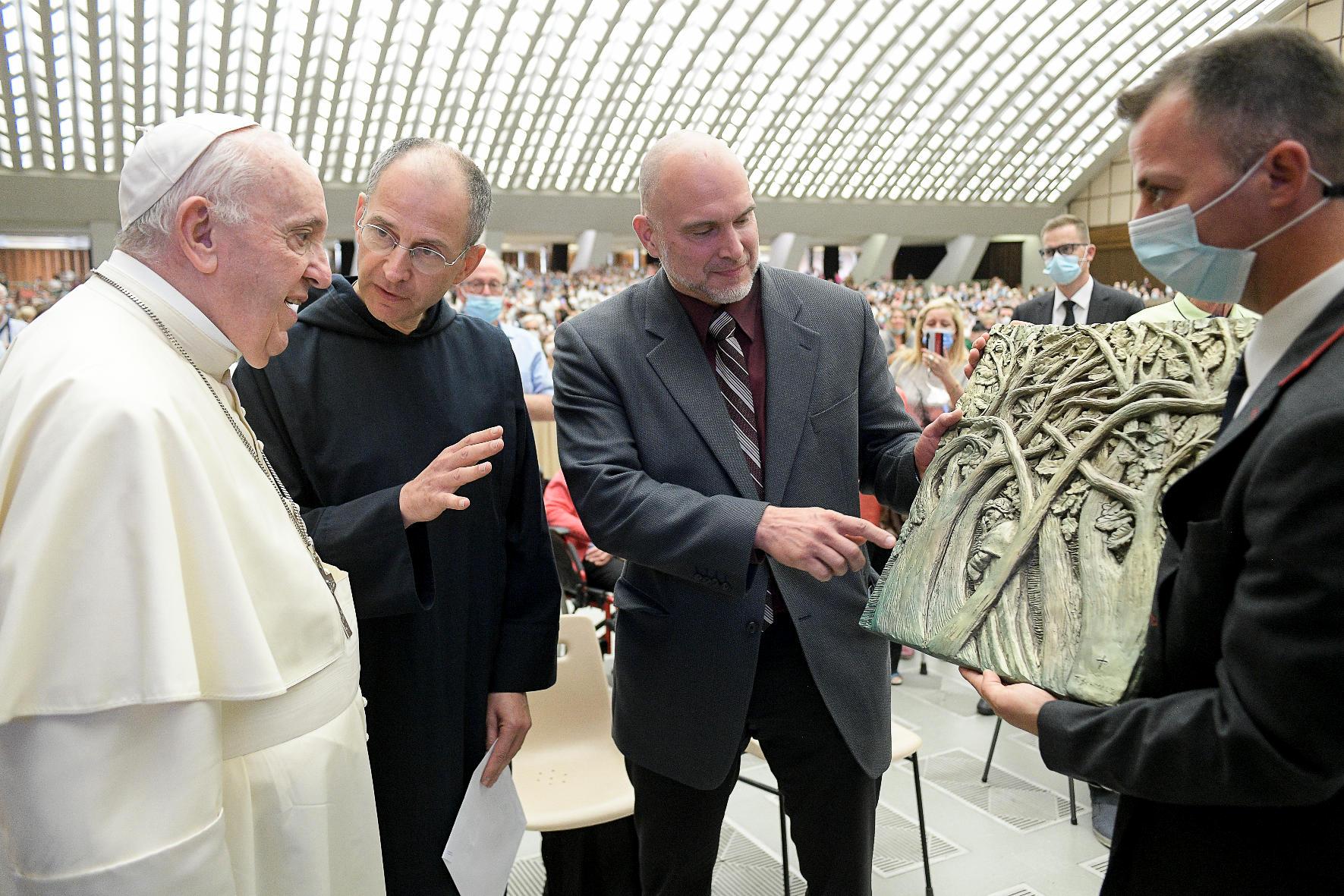 Men in suits hold a sculpted panel depicting intertwining trees up as another man appears to explain the panel to Pope Francis, indoors 