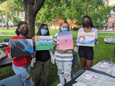 Students wearing masks outdoors and holding paintings next to tables covered in newspaper and painting implements