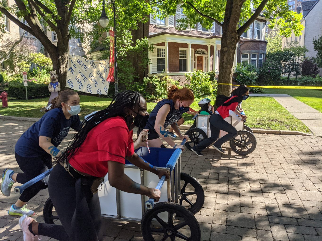 Photograph of the St. Mike's bed race on Elmsley Lane: two teams of three push wheeled carts with a fourth, helmeted team member sitting inside