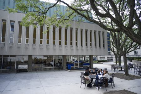 Students sit together at an outdoor table under trees and next to a large campus building 