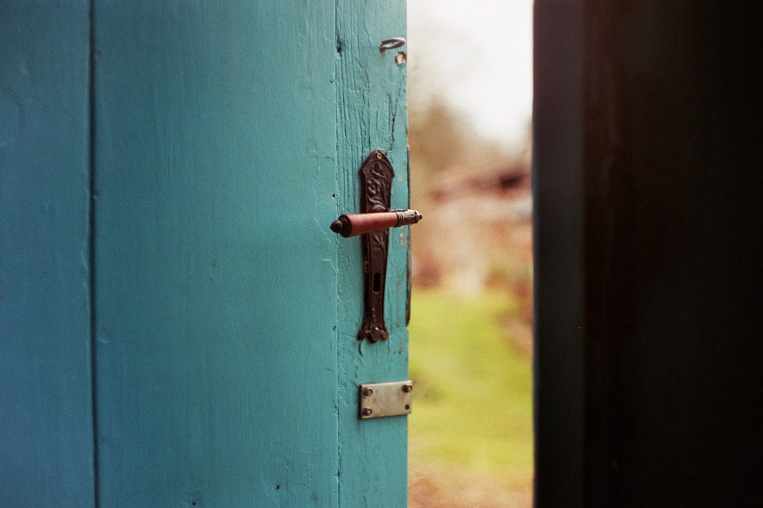 Photograph of an ornate handle on a teal painted wooden door, opened slightly to a green natural background.