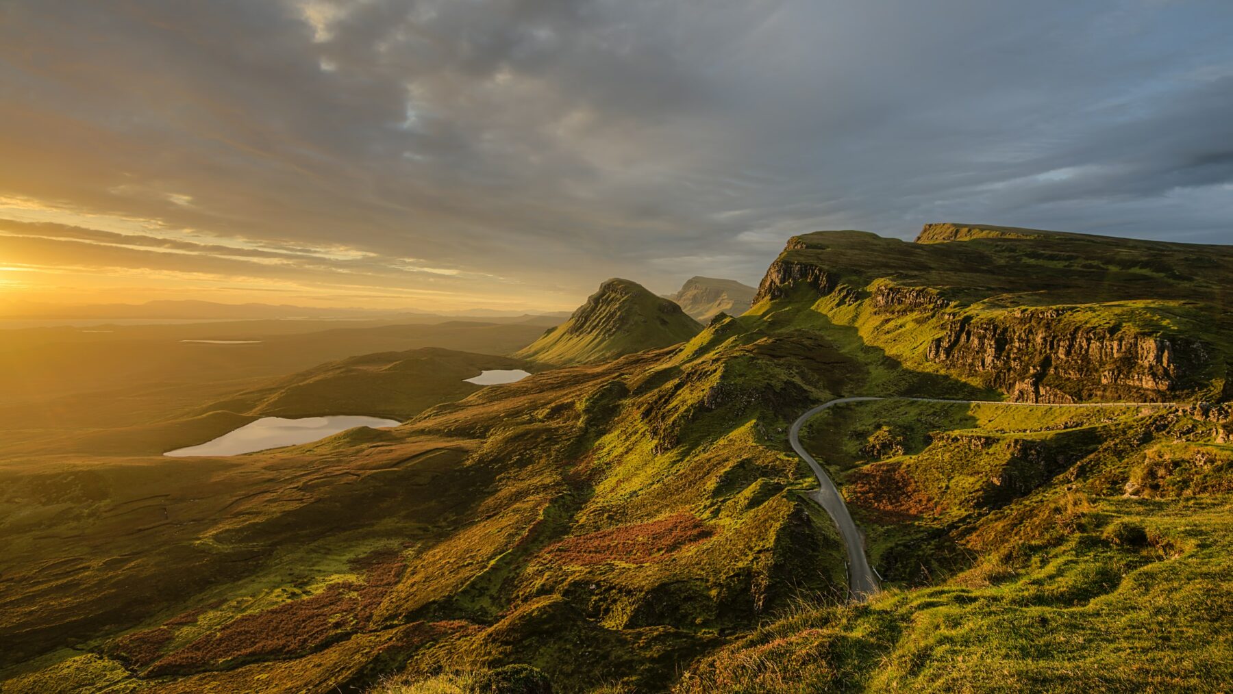 Photograph of a sunset over the mountains of Skye, Scotland, with a winding road in the middle and a swath of clouds in the sky