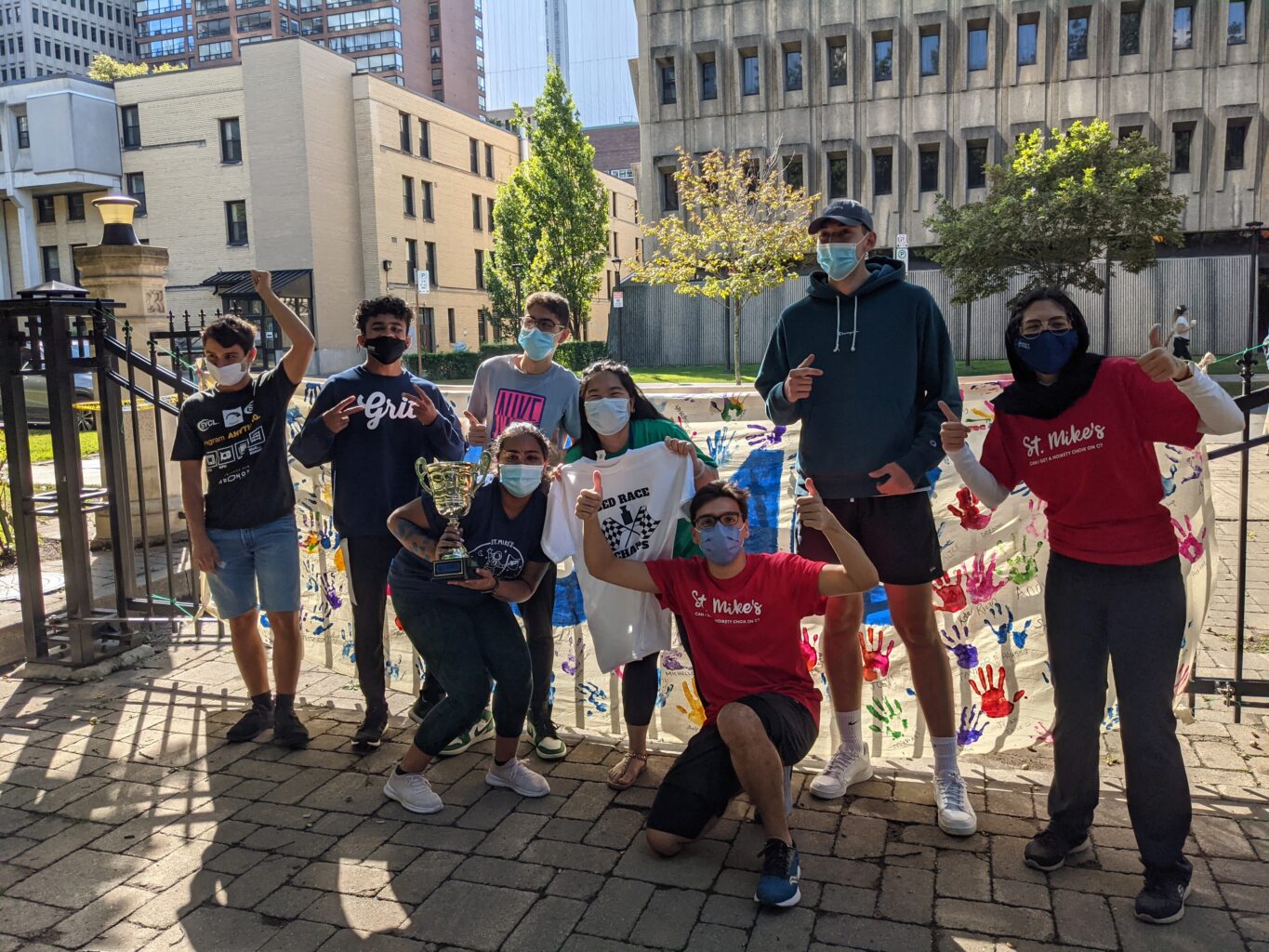 Photograph of seven masked St. Mike's students celebrating with a trophy in front of the gates to Elmsley Place with Kelly Library in the background.