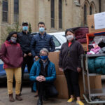 A group of students pose outside St. Basil's on a cold day with Fr. Morgan Rice. Everyone is wearing medical masks and winter coats. They are surrounded by boxes of donations.
