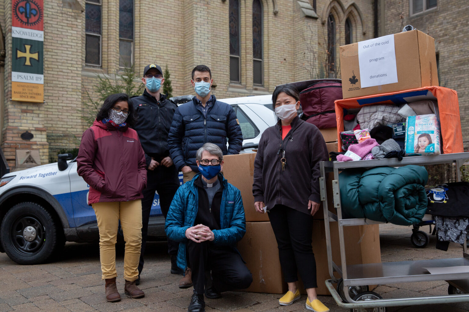 A group of students pose outside St. Basil's on a cold day with Fr. Morgan Rice. Everyone is wearing medical masks and winter coats. They are surrounded by boxes of donations.