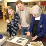 Three people examine archival documents and photos on a table indoors
